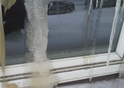 Large, thick icicles with a milky, cloudy appearance hang down a window frame above snow, hinting at potential roofing or siding concerns for Woodbury homes, with cars parked on a snowy street visible in the background.