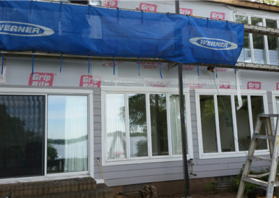 A house in Woodbury under renovation with new siding, large windows reflecting trees and a lake, a blue Werner tarp above, workers on scaffolding, and a ladder in front.