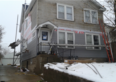 A two-story house in Woodbury with partially installed new siding, construction materials, and ladders around it. Snow covers the ground and sidewalk, while scaffolding is set up along the side—showcasing active general contracting work.