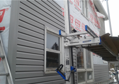 A house under construction in Woodbury with gray vinyl siding partially installed, some walls wrapped in Grip-Rite House-Wrap. General contracting equipment like a ladder and scaffolding is set up alongside the building. Snow covers the ground.
