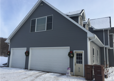 A gray house in Woodbury with siding and a three-car garage is decorated with holiday wreaths. Snow covers the ground and roofing. Three brown garbage bins and a bag of salt sit near a side door under a partly cloudy sky.