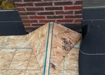 A piece of plywood, cut and shaped like a paper airplane, lies on a plywood floor in front of a brick-patterned wall—its smooth surface reminiscent of the quality found in Woodbury roofing projects. Various objects and cables are partially visible nearby.