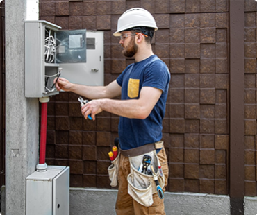 An electrician wearing a hard hat and safety glasses works on an outdoor electrical panel, using tools from his belt. The panel is mounted on a post beside a textured brown wall with new siding installed by a general contracting team.