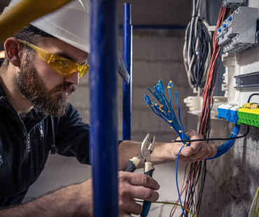 An electrician wearing a hard hat and safety glasses uses wire cutters to work on electrical wiring inside an electrical panel mounted on a wall at a general contracting site. Multiple colored wires are visible.