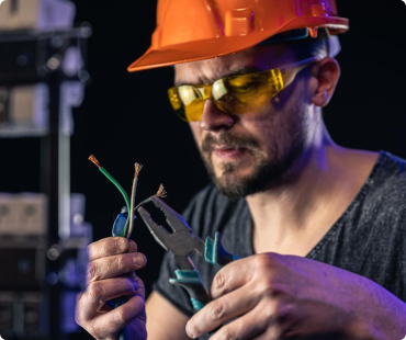 A man wearing an orange hard hat and yellow safety glasses uses pliers to work on exposed electrical wires while repairing siding in Woodbury, holding them carefully in his hands.