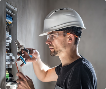 A man wearing a white hard hat, safety glasses, and a black t-shirt uses pliers to work on an electrical panel mounted on a wall, showcasing his skill in general contracting tasks.