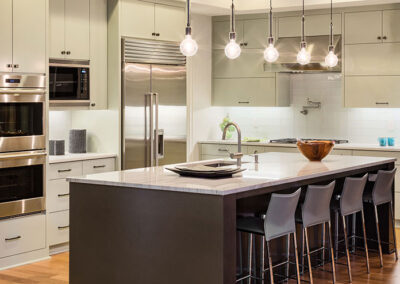 Modern kitchen with a large island, gray cabinets, stainless steel appliances, and pendant lights. Four barstools face the marble countertop, while decorative items sit on the island—a stunning space in Woodbury perfect for entertaining.