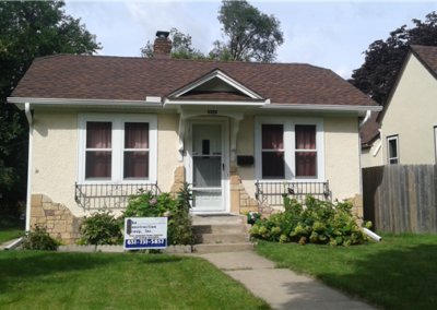 A small, light-yellow house with a brown roof features six front windows and a white door. A General Contracting sign is in the yard, surrounded by green plants, highlighting quality craftsmanship in the heart of Woodbury.