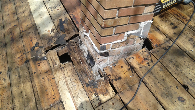 A damaged wooden deck surrounds a brick chimney, with siding near the base showing signs of rot. Large holes and dark stains mar the wood, while a power tool and extension cord rest on the deck nearby.