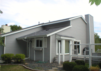 A light gray, single-story house with modern, angular architecture, white trim, new siding, a sloped roof, large windows, a small deck, and landscaped shrubs in the yard under a partly cloudy sky.