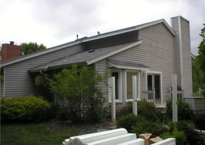 A modern, light gray house with angular rooflines and white trim in Woodbury, surrounded by green bushes and trees. Gutters and building materials are stacked in the yard, suggesting ongoing construction or general contracting work.
