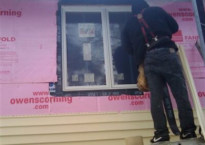 A person stands on a ladder in Woodbury, installing or working on a window of a house under construction, with pink Owens Corning insulation, yellow siding, and new gutters visible.