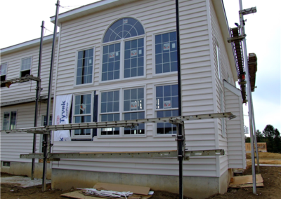 A white house under construction features large, multi-pane windows and exterior scaffolding. The ground is bare dirt, with building materials scattered nearby—a typical scene on a General Contracting job site.