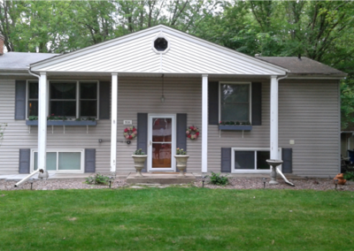 A light gray split-level house with white trim, four columns on the porch, and flower pots near the front door. The yard has green grass, new windows, and gutters installed by a trusted general contracting team. Trees surround the house.