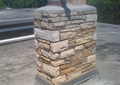 A stone chimney with two white PVC pipes stands on a flat, gray rooftop with expertly installed gutters, all under a clear blue sky and framed by green trees in the background.