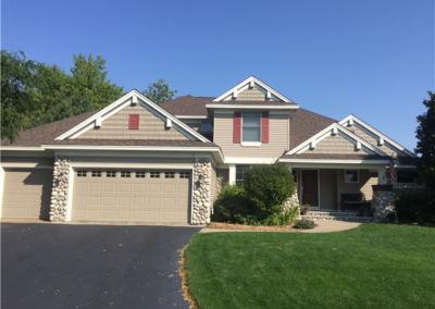 A two-story suburban house in Woodbury with a stone-accented exterior, three-car garage, neatly trimmed lawn, and a paved driveway under a clear blue sky. Gutters and large windows enhance the home, while shrubs and trees add greenery.
