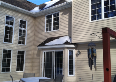 A beige house in Woodbury with siding, tall windows, and a glass patio door, partially covered with snow. Patio furniture and a wind chime hang nearby, with snow visible on the roof and ground.