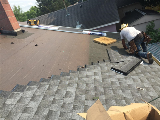 A worker in a hat and tool belt installs gray shingles on a sloped roof in Woodbury, with tools, roofing materials, and nearby windows gleaming under a sunny sky.
