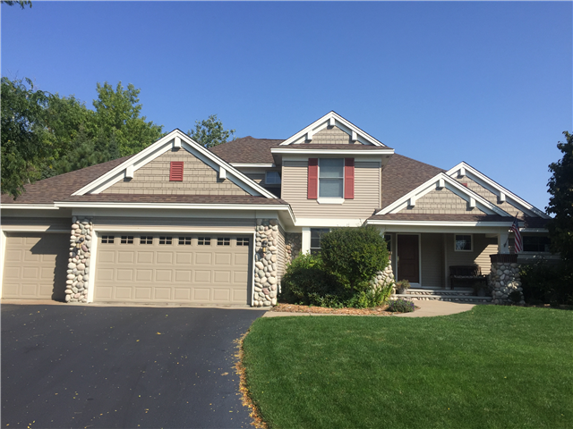 A modern suburban house in Woodbury with tan siding, stone accents, gutters, a three-car garage, manicured lawn, shrubs, and trees under a clear blue sky. An American flag hangs near the front porch.