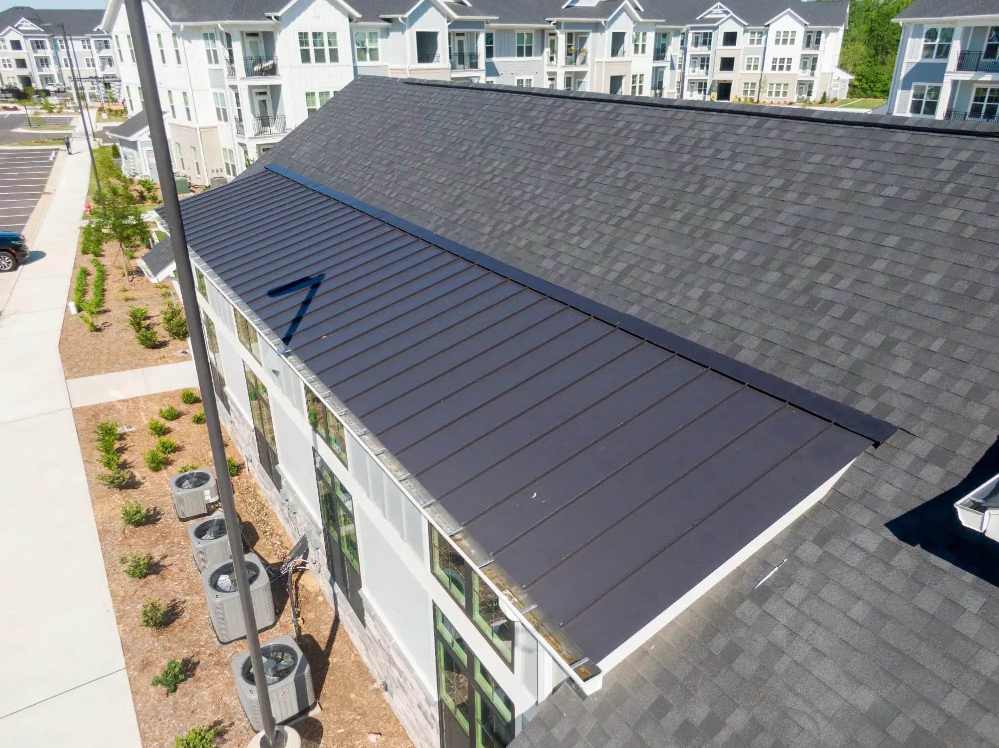 Aerial view of a building with a black metal roof overhang and dark shingles, surrounded by modern multi-story apartment buildings and landscaped areas.
