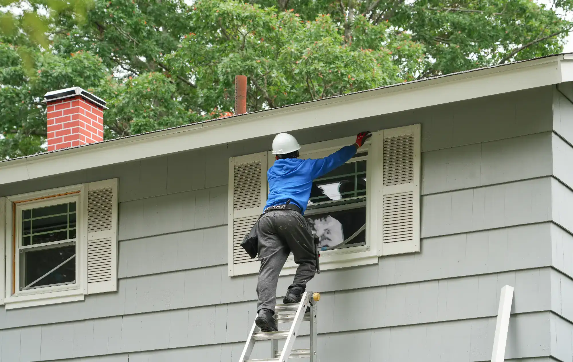 A worker in a blue shirt and white helmet stands on a ladder, installing or repairing a window on the second floor of a gray house with white trim and surrounded by green trees.