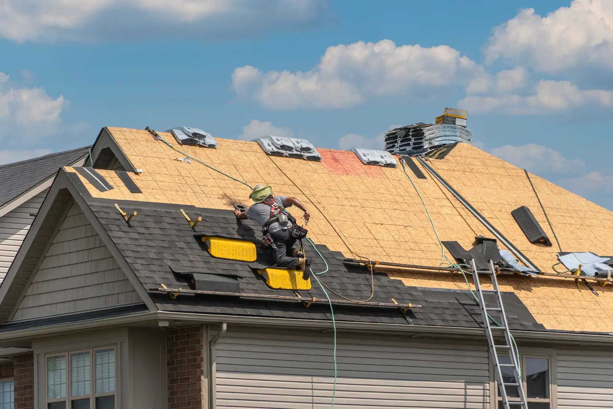 A worker wearing safety gear repairs the roof of a house, installing new shingles. Construction materials and tools are scattered on the partially completed roof under a blue sky with clouds.