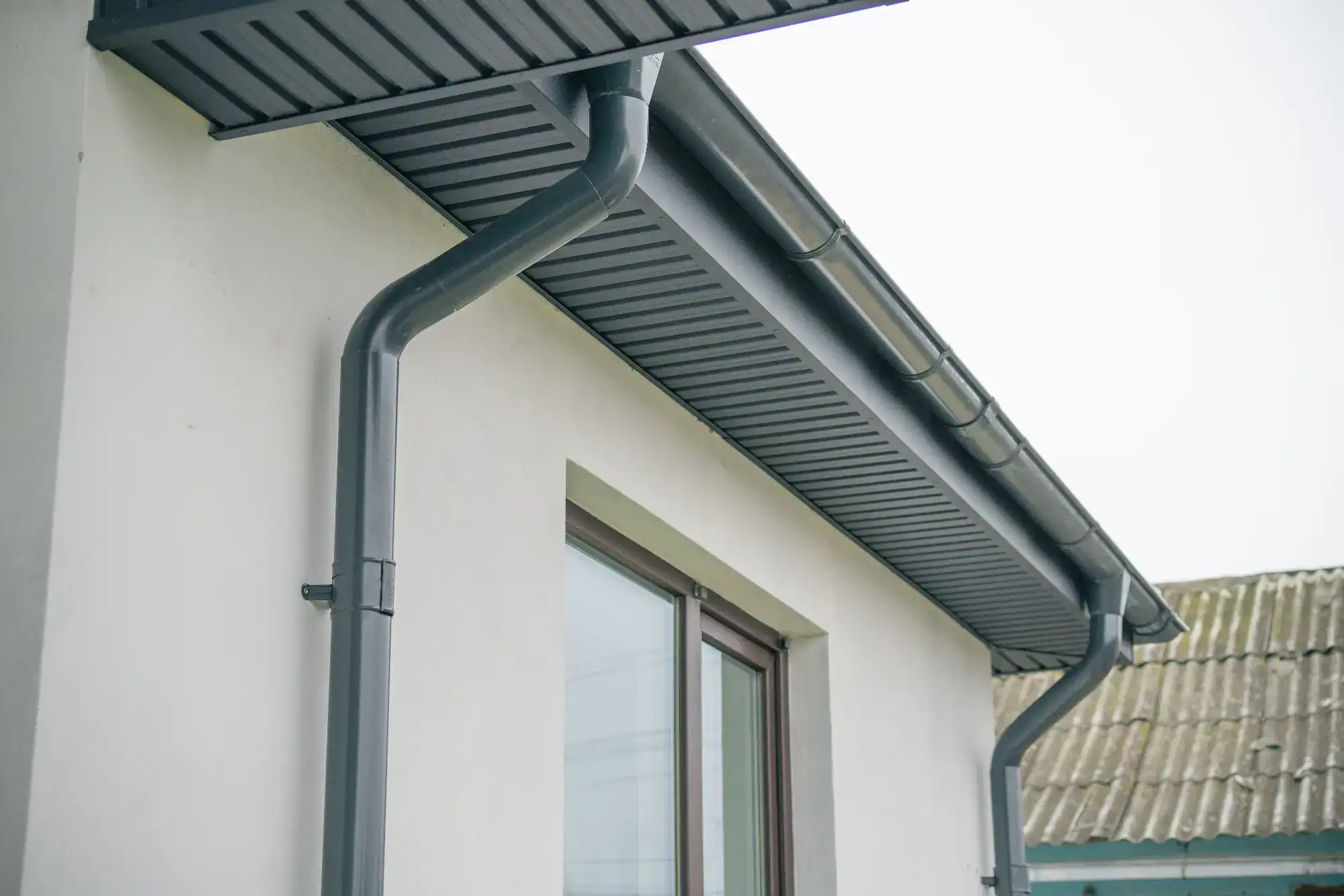 Close-up view of the side of a house showing a gray metal gutter and downspout system attached to the roof, with a window below and part of another roof visible in the background.
