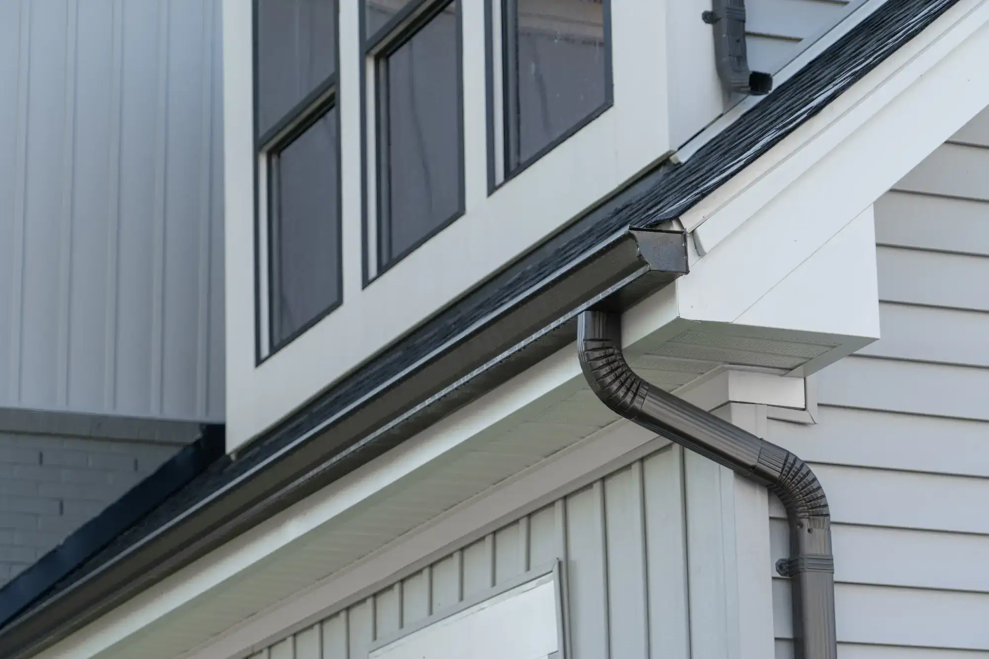 A close-up view of a house exterior showing gray siding, a black gutter and downspout, and several large windows reflecting the sky.