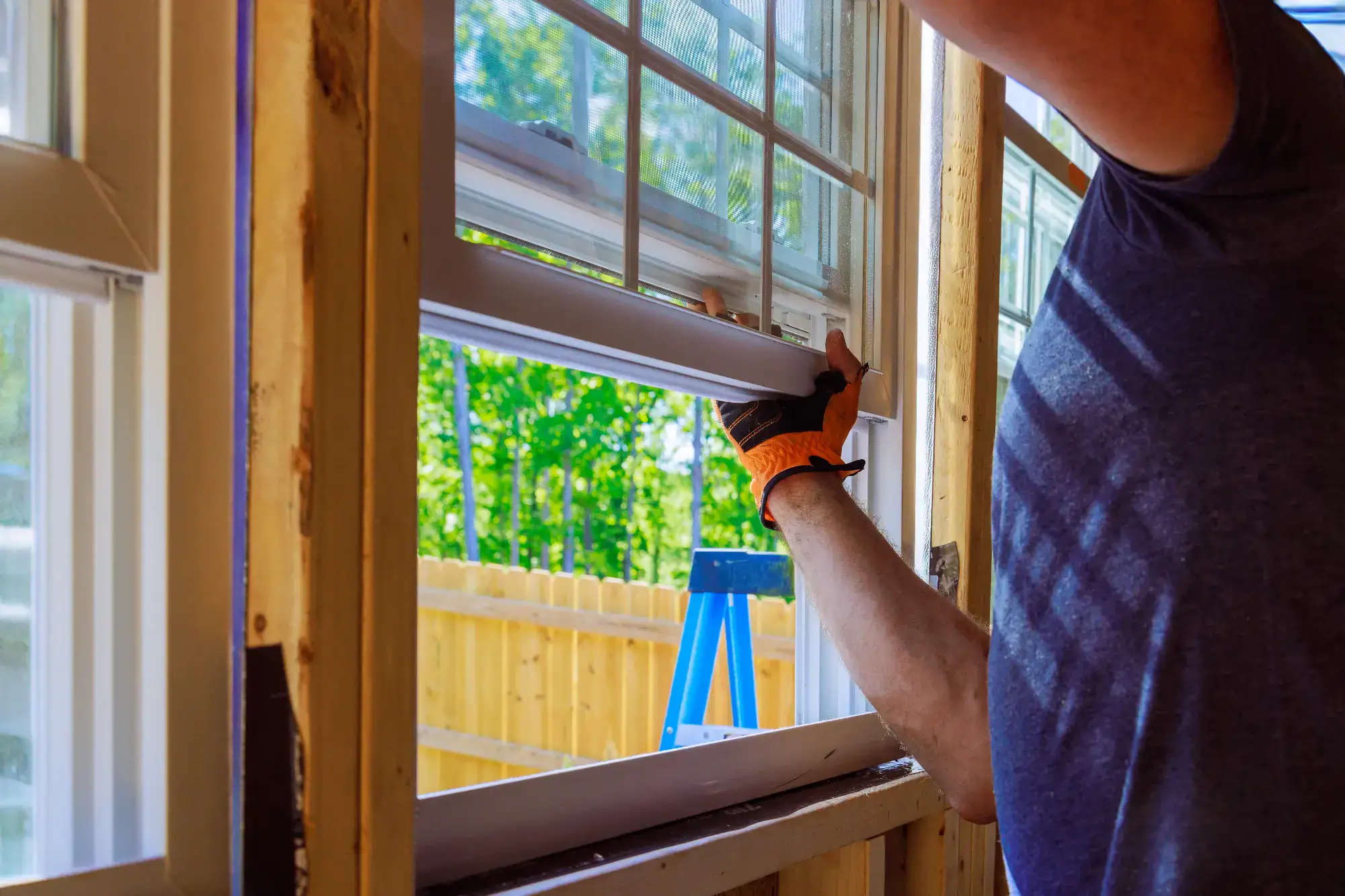 A person wearing orange gloves installs a window in a house under renovation, with a wooden fence and green trees visible outside.