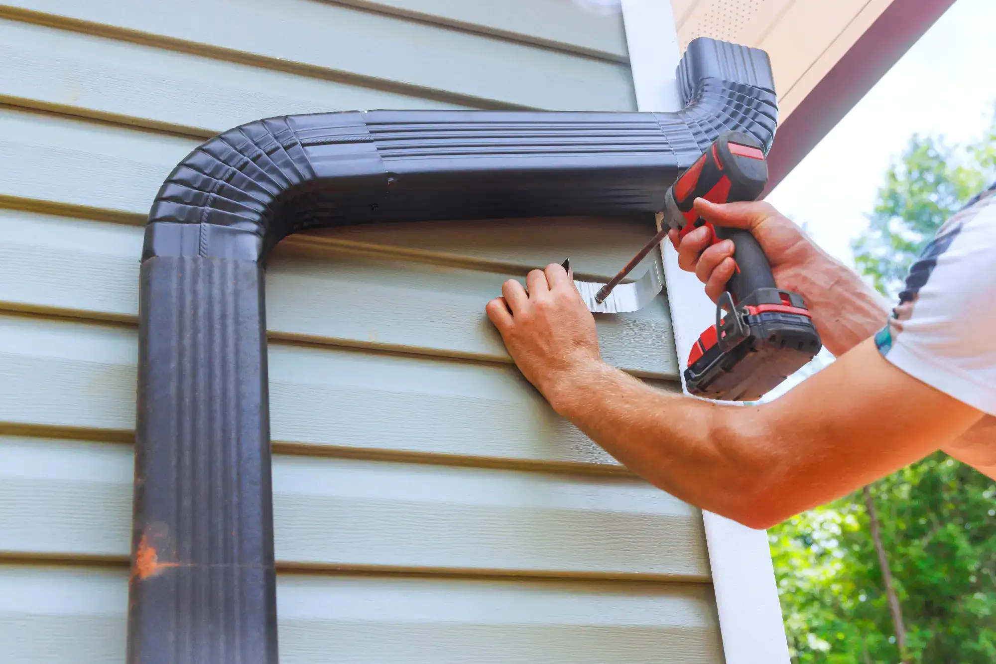 A person uses a power drill to attach a black rain gutter downspout elbow to the exterior wall of a house with light blue siding.