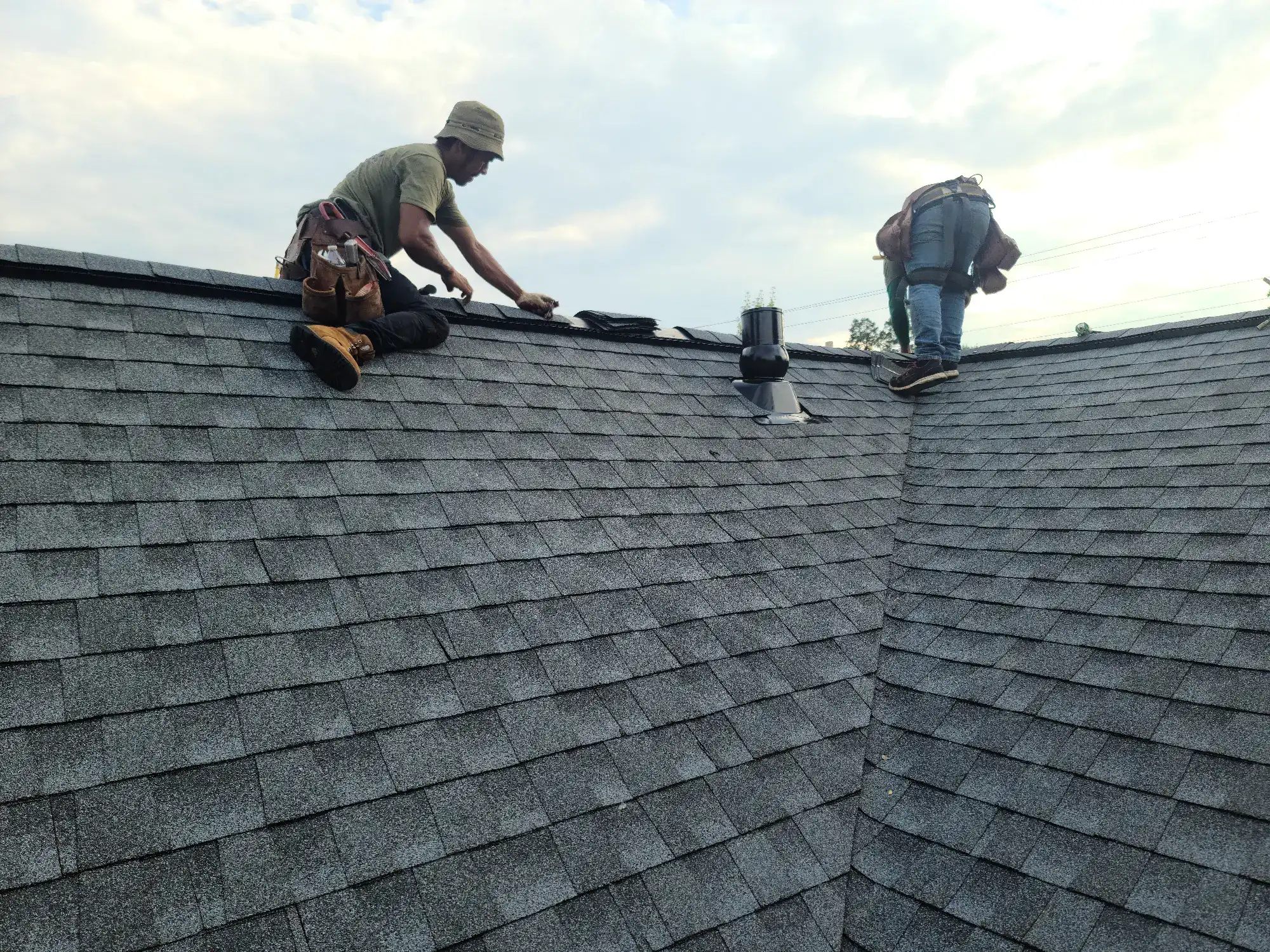 Two workers wearing tool belts are installing or repairing shingles on the roof of a house under a partly cloudy sky. One is sitting while the other is bent over, both focused on their tasks near the ridge of the roof.