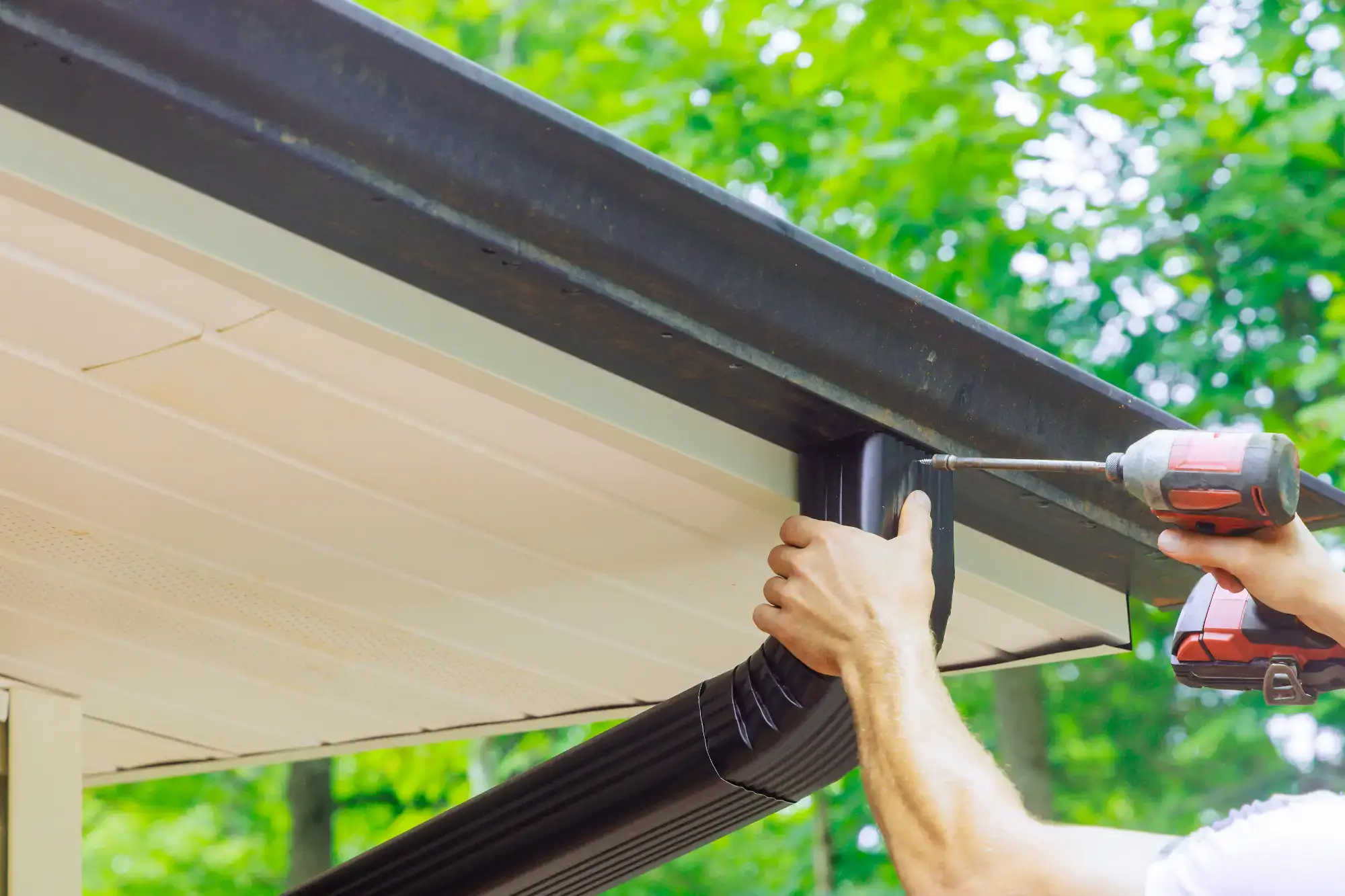 A person uses a power drill to attach a black downspout to the gutter of a house, with green trees visible in the background.