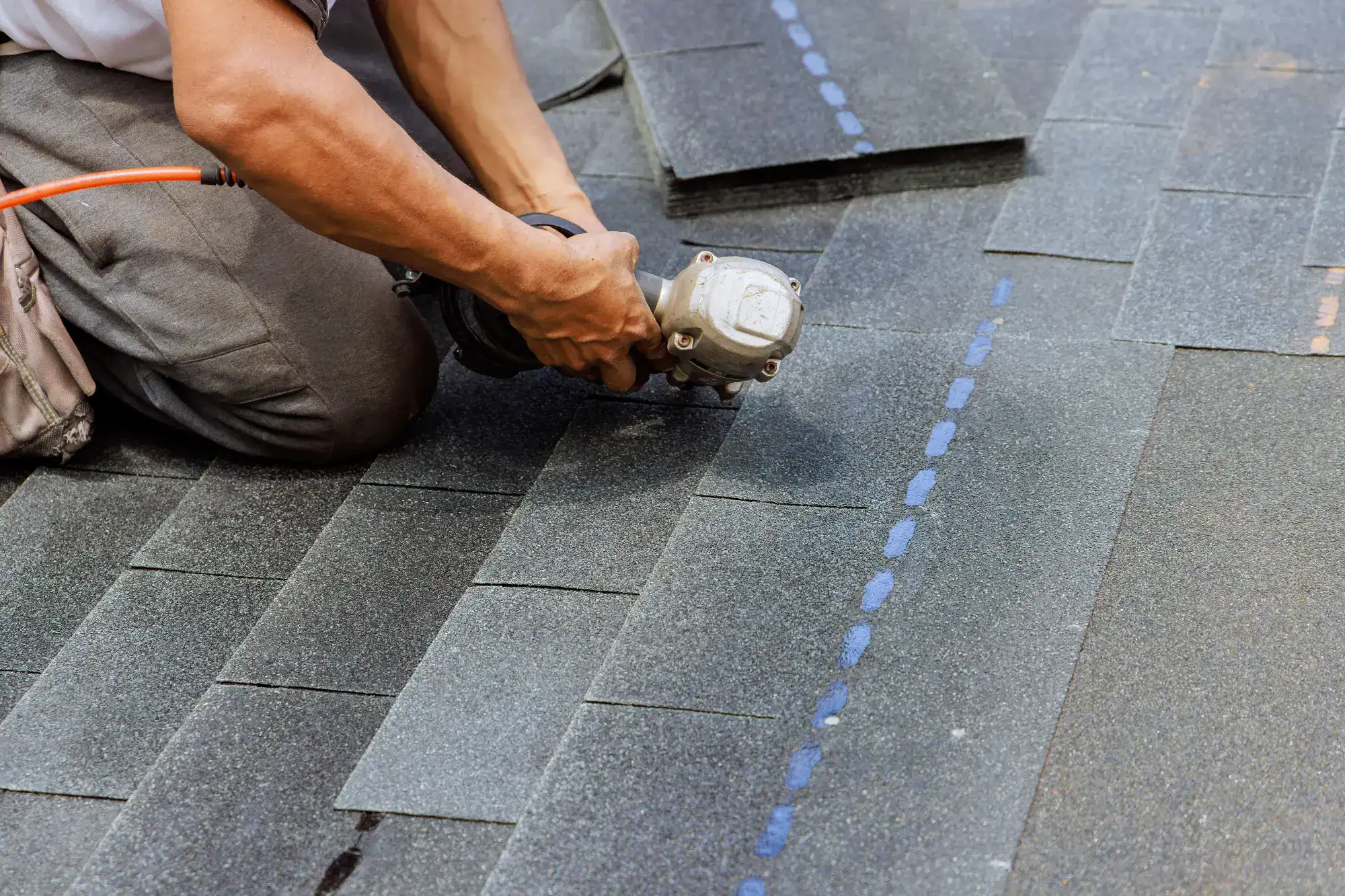 A person kneeling on a roof uses a power tool to fasten dark asphalt shingles, installing roofing material in overlapping rows.