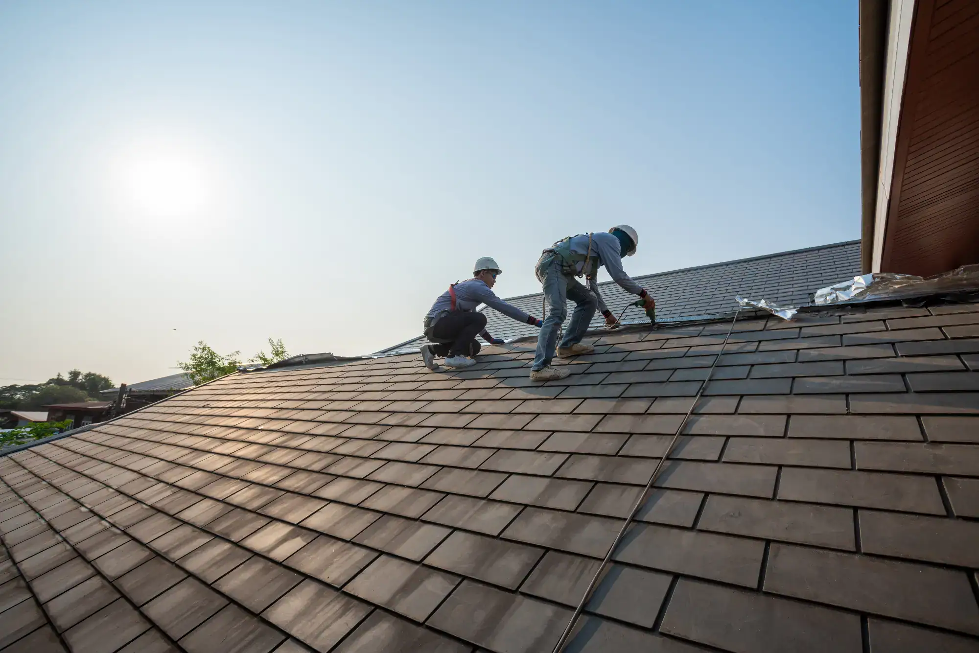 Two construction workers wearing safety gear work on a sloped roof covered with shingles under a clear sky, with sunlight shining in the background.