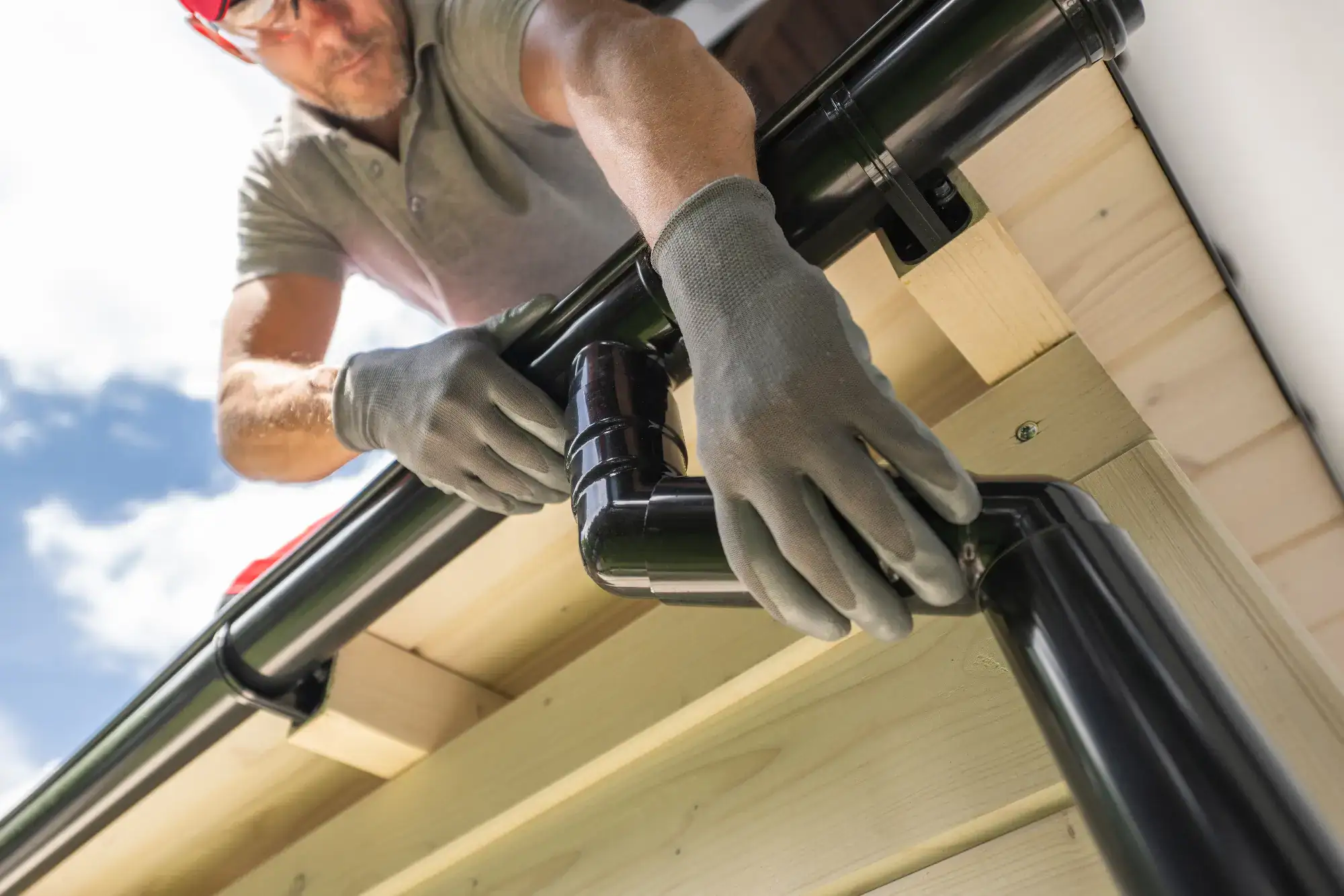 A person wearing gloves installs a black rain gutter and downspout on the edge of a wooden roof, working outdoors on a sunny day.