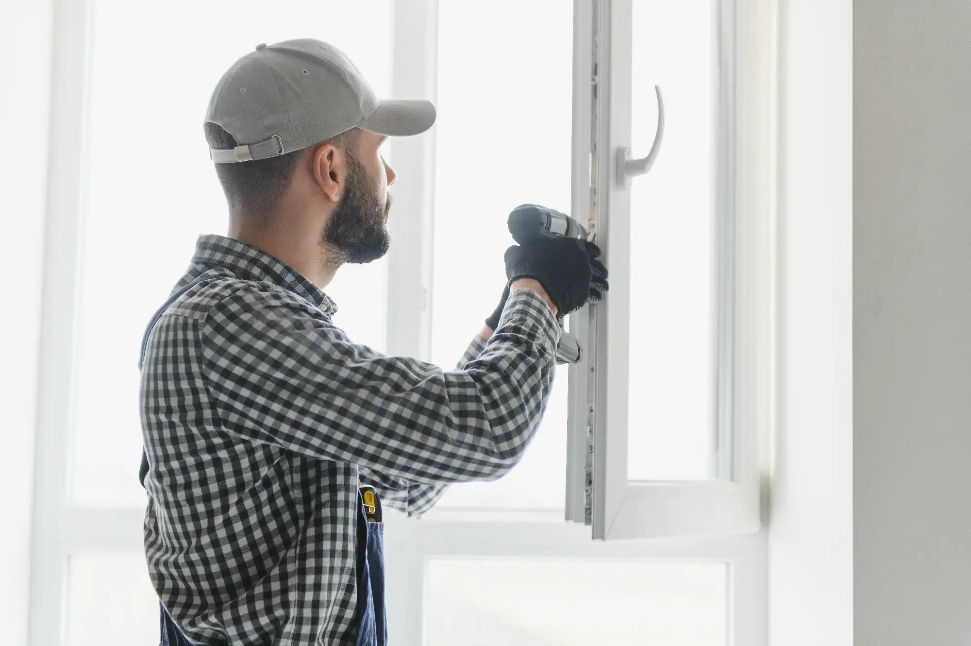 A man wearing a checkered shirt, cap, and gloves uses a power drill to install or repair a white window in a bright, sunlit room.