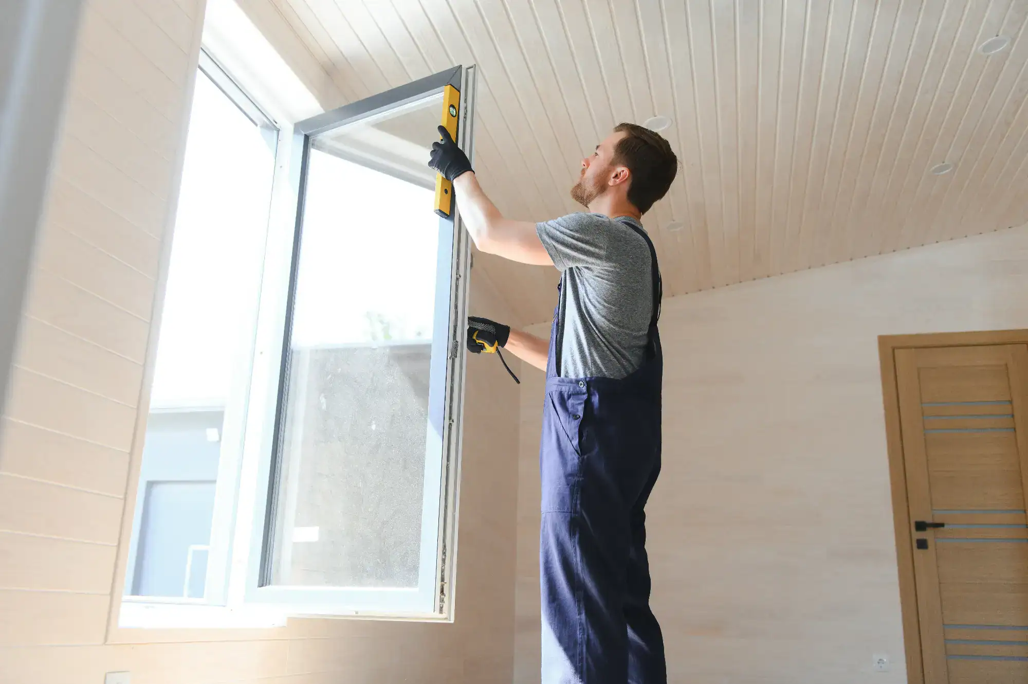 A man wearing work overalls and gloves uses a level tool to adjust or install a window frame in a brightly lit room with wooden walls and ceiling.